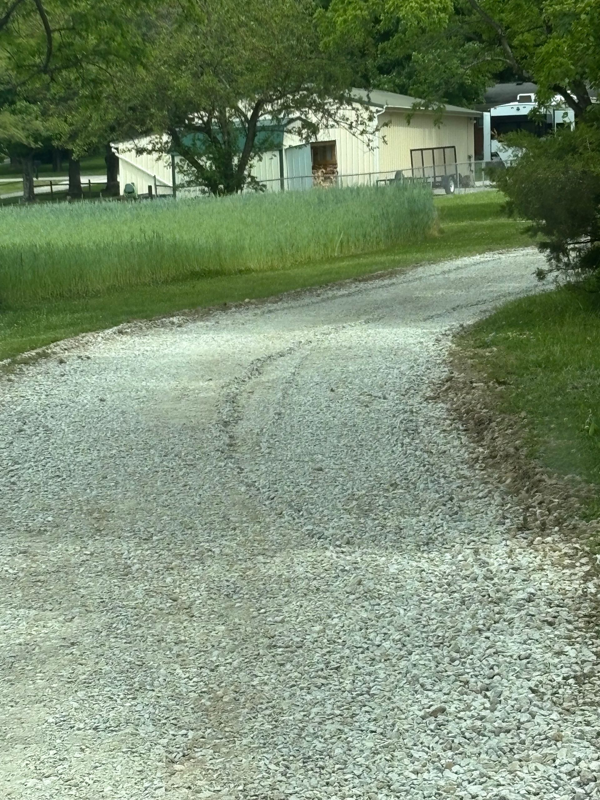Gravel driveway curving past a grassy field toward a small house and parked vehicle.
