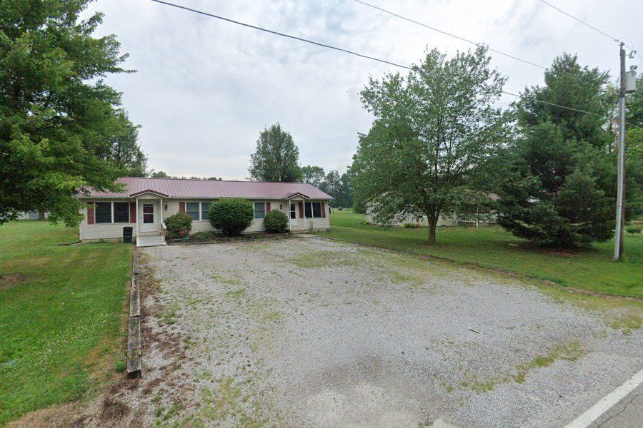 Single-story house with a gravel driveway and trees under a cloudy sky.