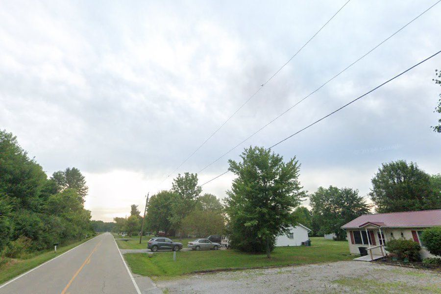 Rural roadside with parked cars, trees, power lines, and a small house under a cloudy sky