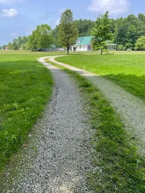 Gravel driveway curving through a green field toward a small house under a partly cloudy sky