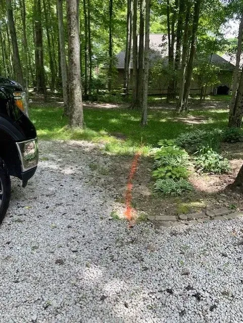 Gravel driveway beside a wooded yard with a black SUV and a house in the background.