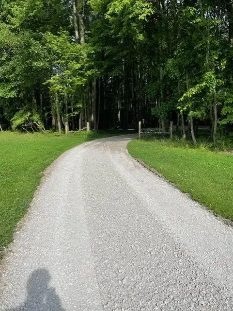 Gravel path curving through a green wooded park under trees, with a shadow in the foreground