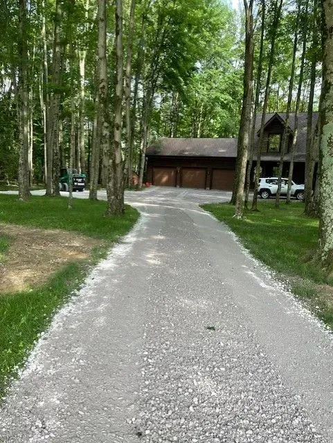 Gravel driveway through tall trees leading to a wooden garage and parked white SUV