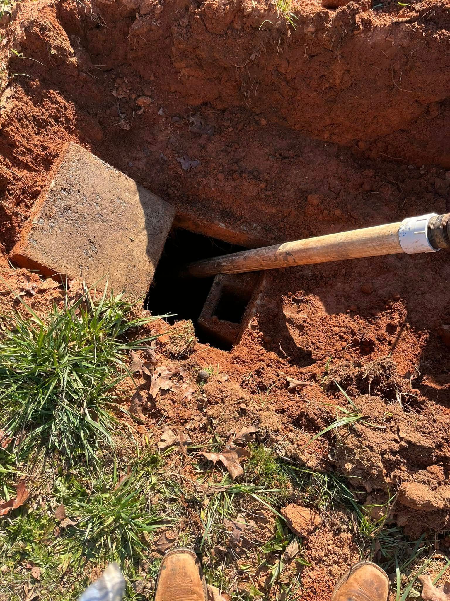 A square hole dug in red soil, revealing a concrete structure. A shovel is inside the hole.
