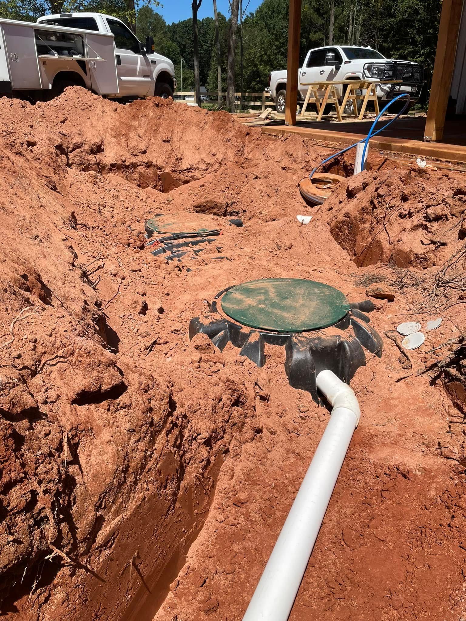 Septic tank installation in progress with exposed pipes and red soil. Construction vehicles in the background.