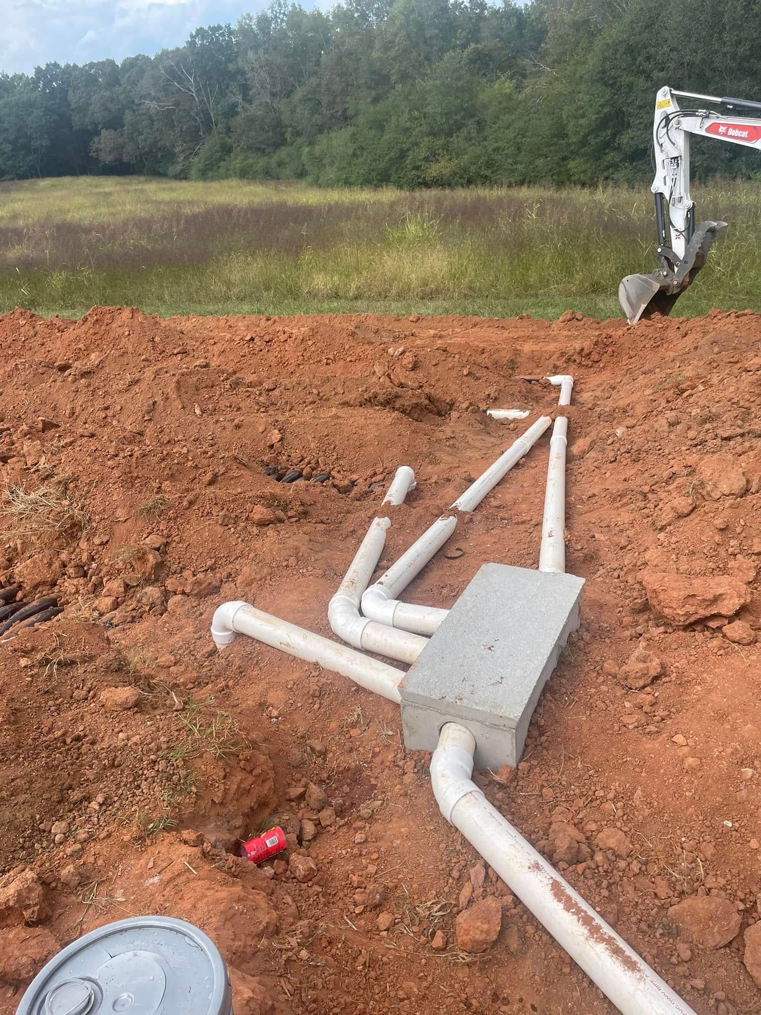 Excavated area with white pipes, a concrete box, and a small excavator. Red dirt with a tree line in the background.