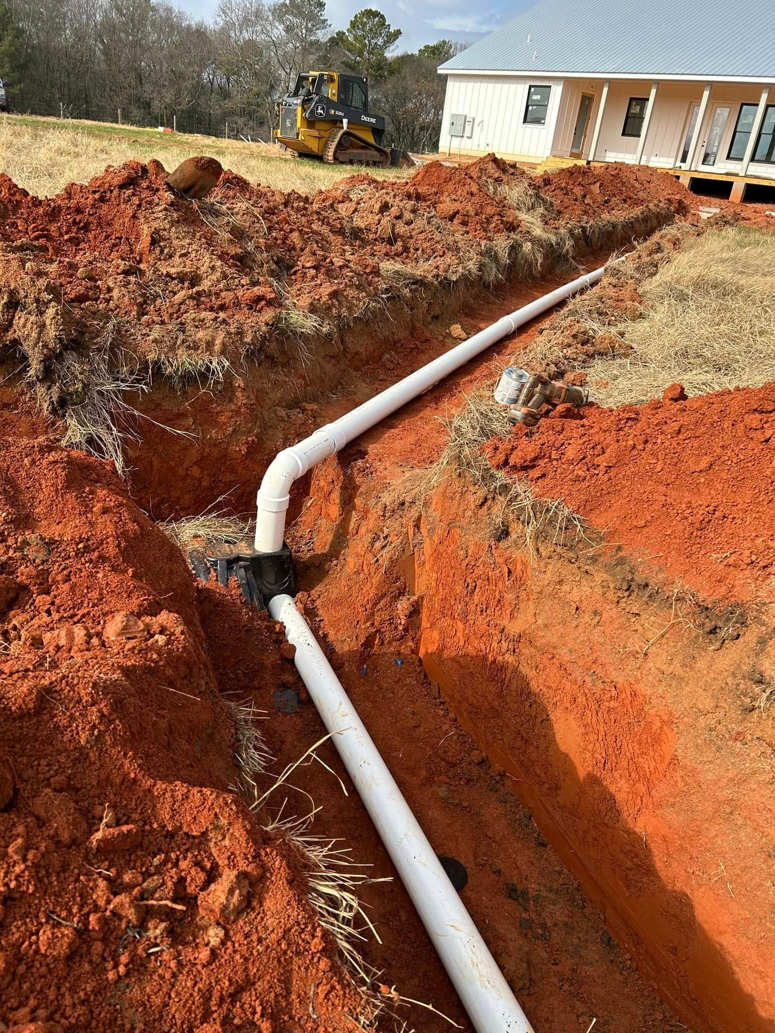 White pipes in a trench for construction; red soil surrounds, with a house and machinery in the background.
