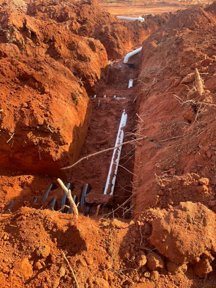 Trench with white and black pipes running through red clay soil, outdoor setting.
