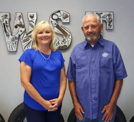 Two people standing indoors in front of wall letters, one in a blue top and one in a blue shirt | Westside Tire & Service