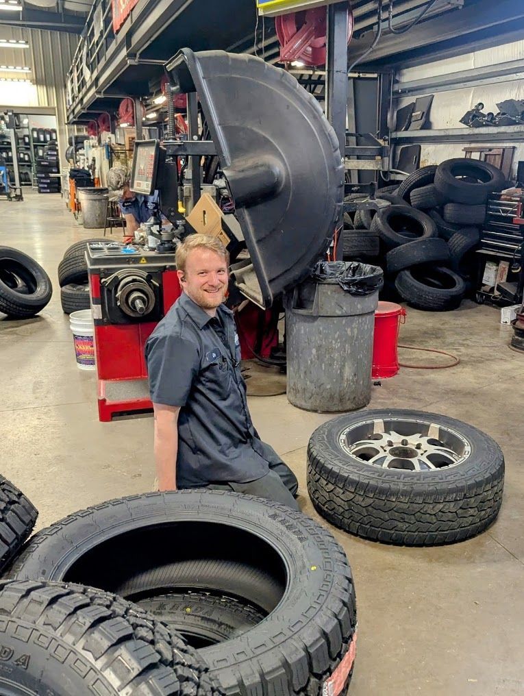 Mechanic kneeling in an auto shop beside stacked tires and a lifted tire machine | Westside Tire & Service