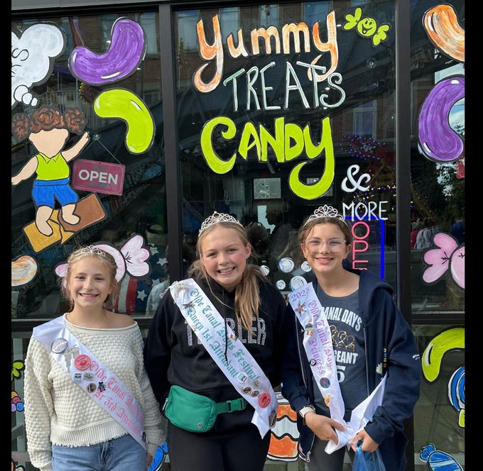 Three girls posing in front of a colorful candy shop window with “Yummy Treats Candy” painted on it.