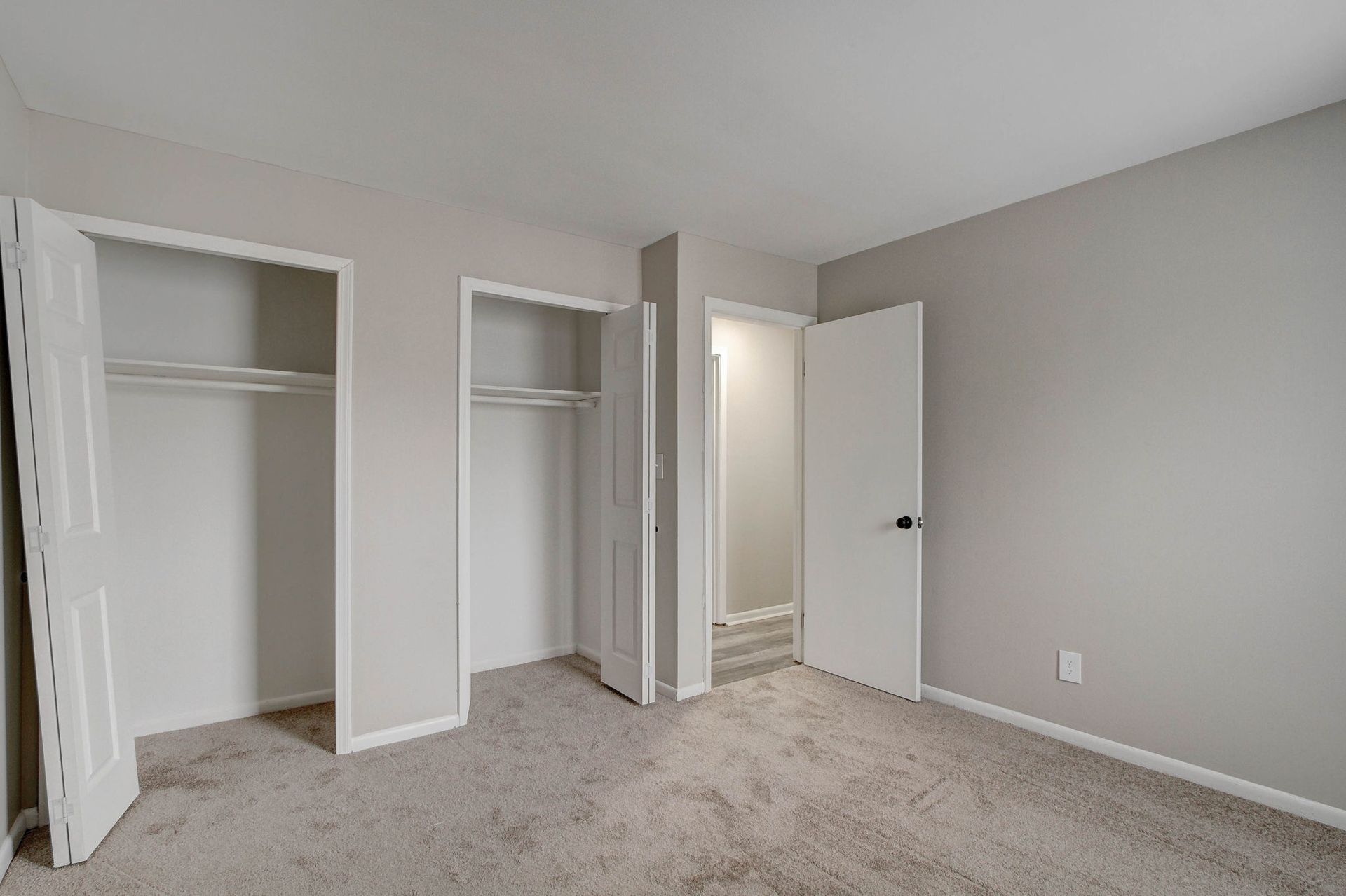 Empty bedroom with two closets and an open door to a hallway. Gray walls and carpet.