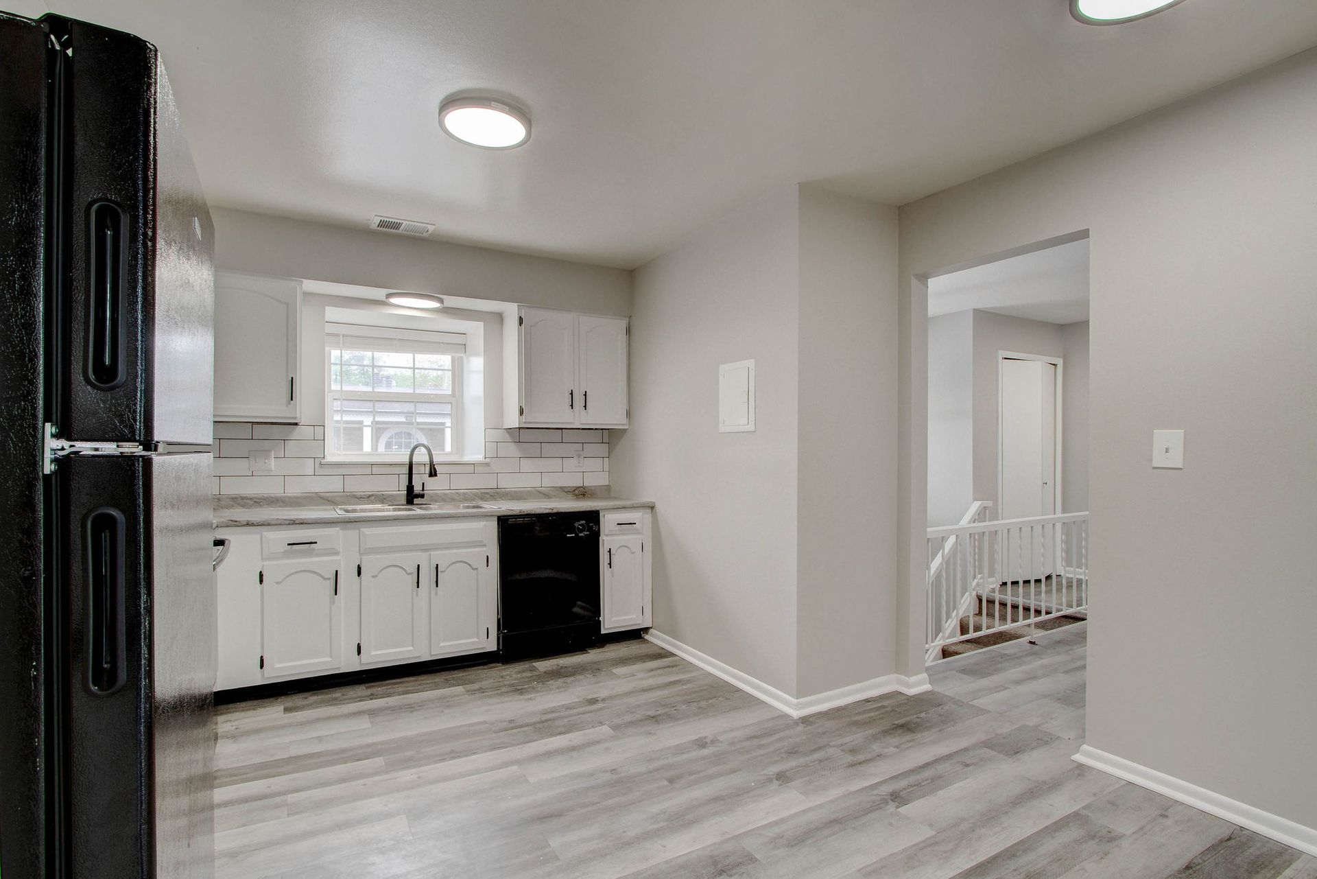 Kitchen with white cabinets, black appliances, and grey flooring. Adjacent hallway with stairs.