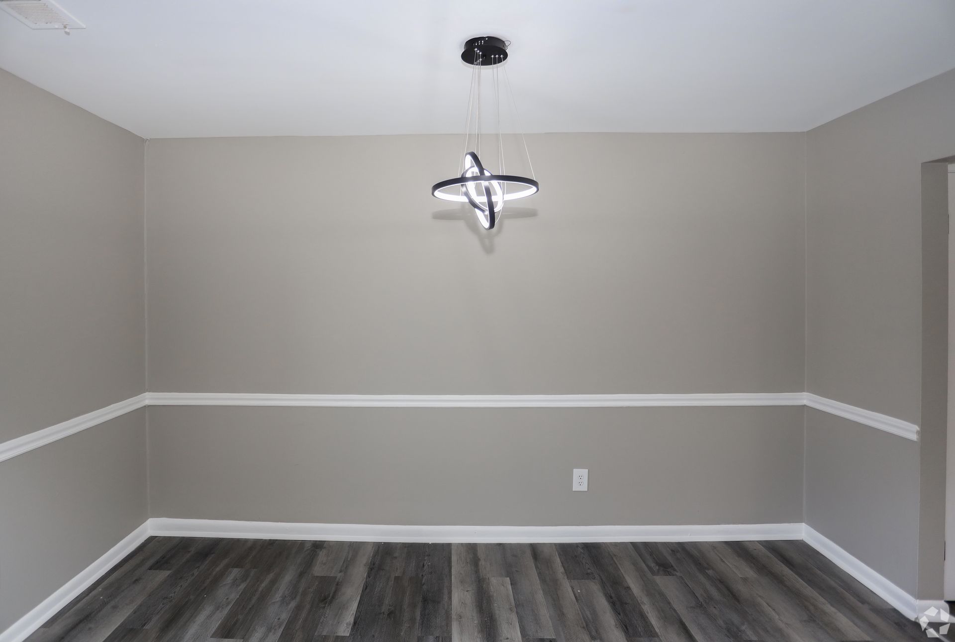 Empty dining room with gray walls, white trim, and modern chandelier. Dark wood-look flooring.