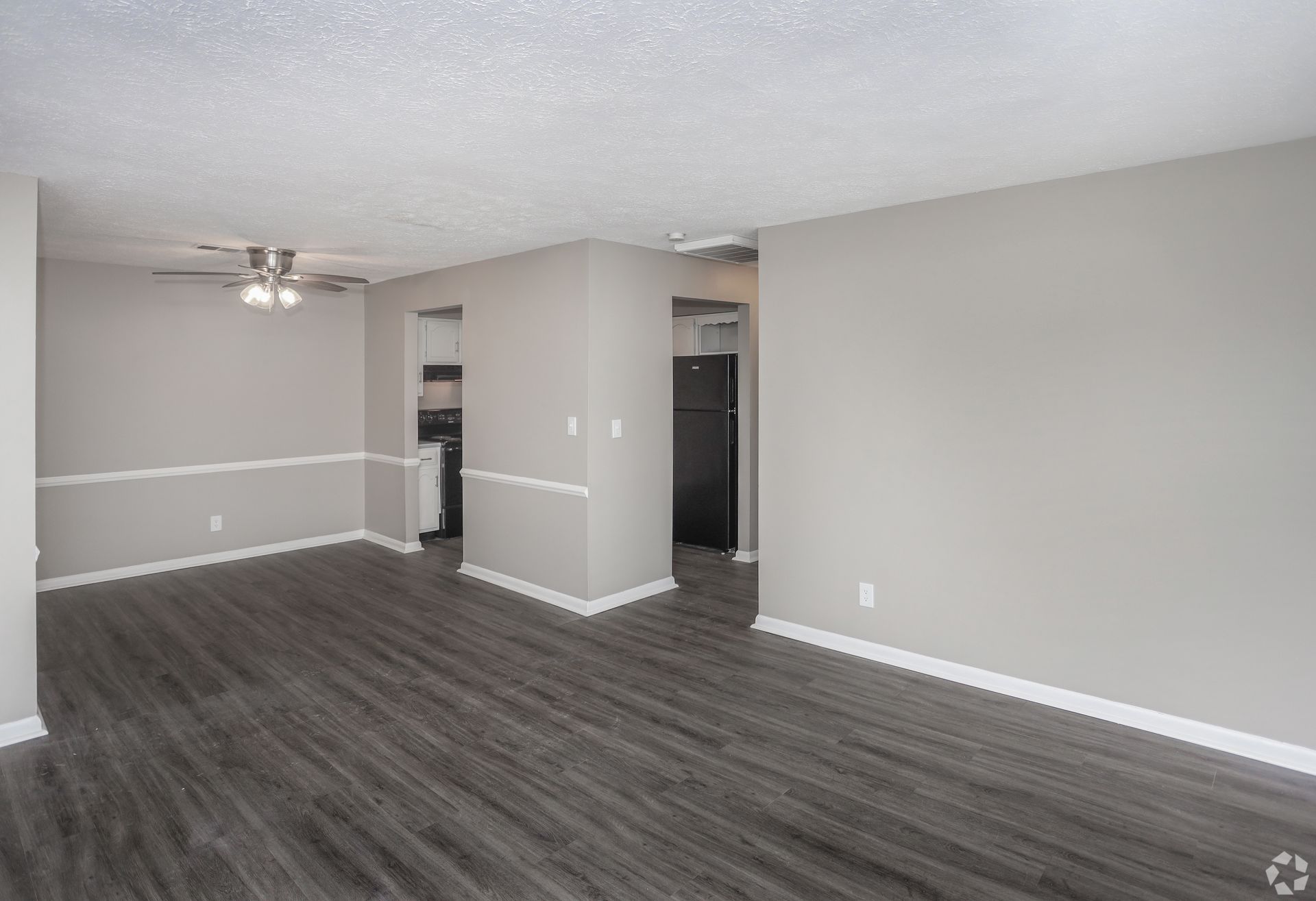 Empty apartment interior with gray walls, flooring, and trim. A refrigerator is visible in the doorway.