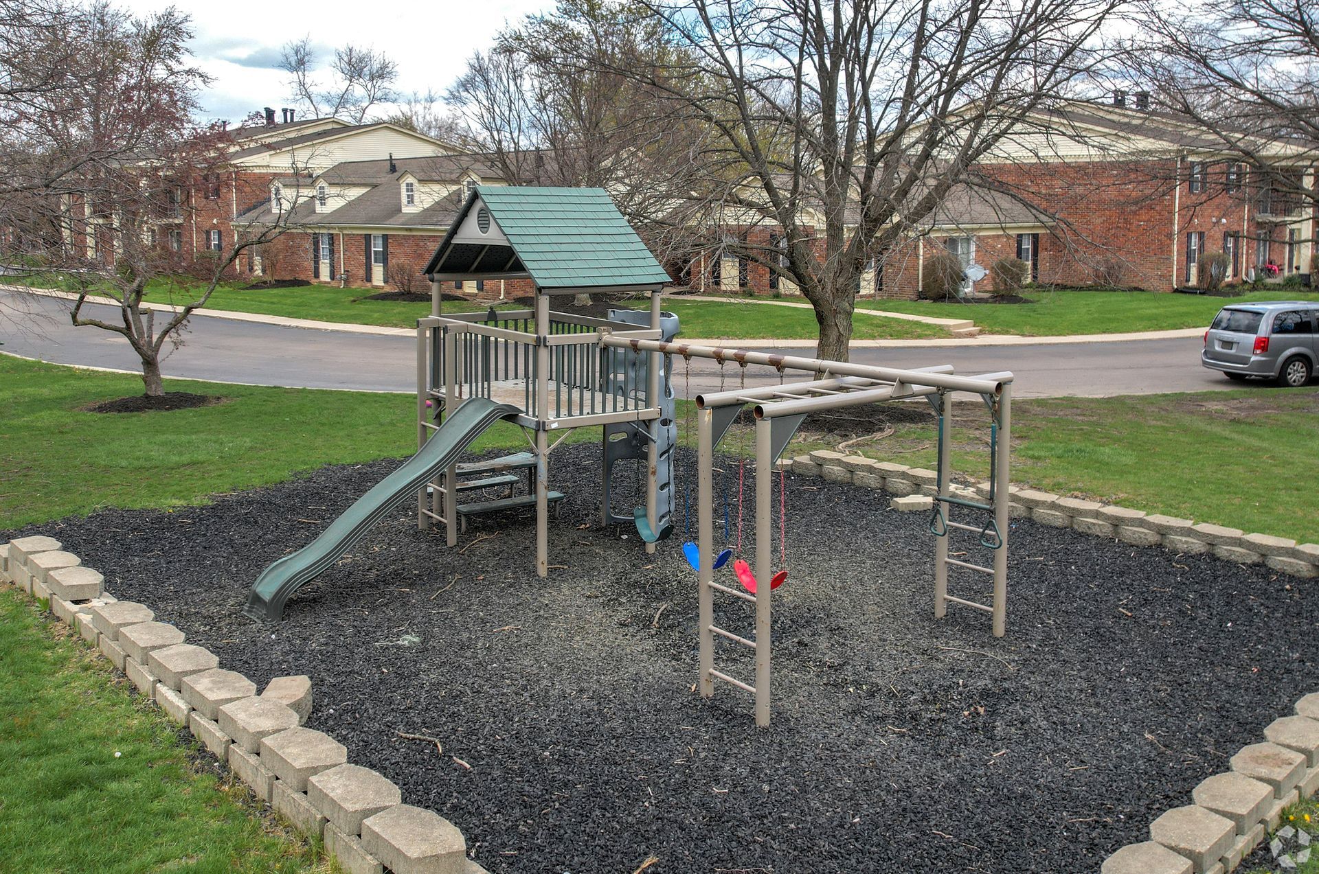 Playground with slide, climbing bars, and black rubber ground in a grassy area near apartment buildings.
