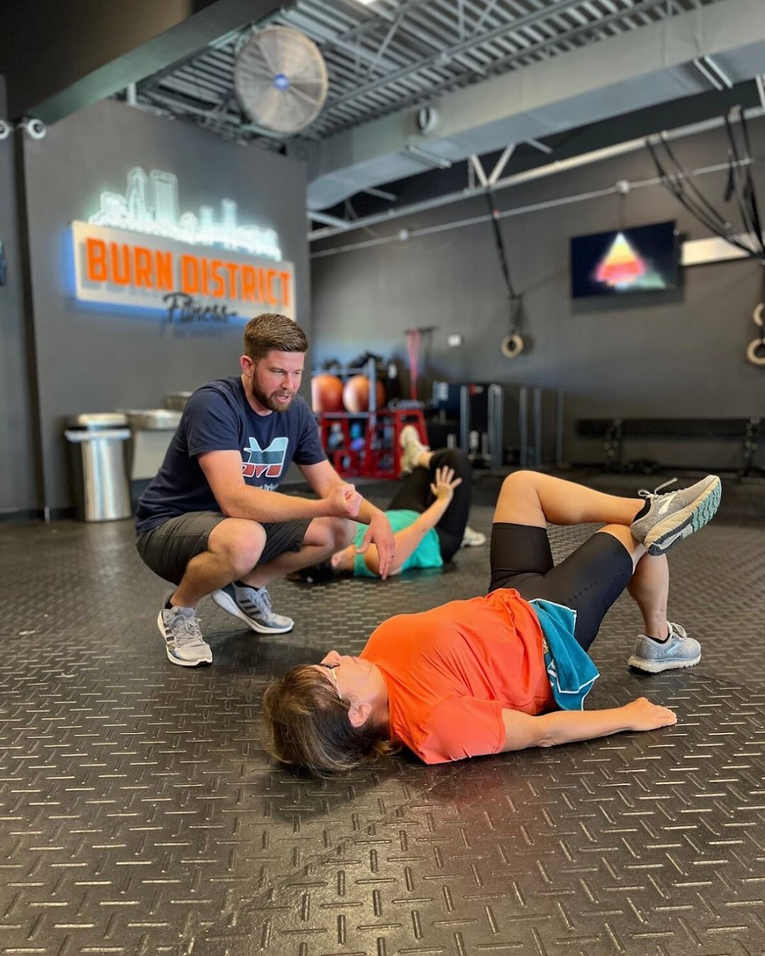 A trainer assists a person performing a glute bridge exercise on a gym floor.
