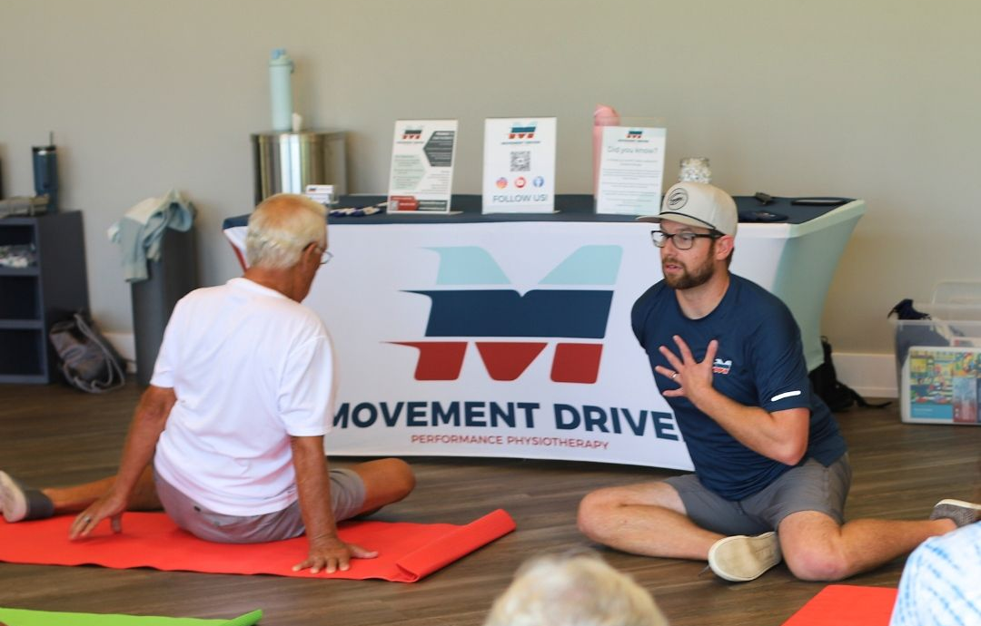 Man instructing seated person on red mat, with Movement Driven logo and products in background. Man instructing seated person on red mat, with Movement Driven logo and products in background.