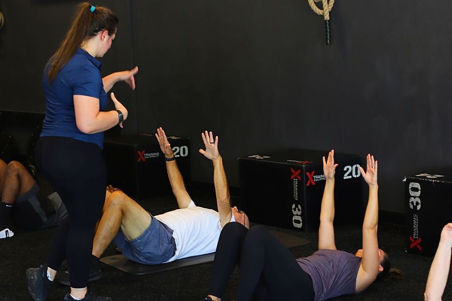 A woman is standing next to a group of people doing exercises in a gym. A woman is standing next to a group of people doing exercises in a gym.