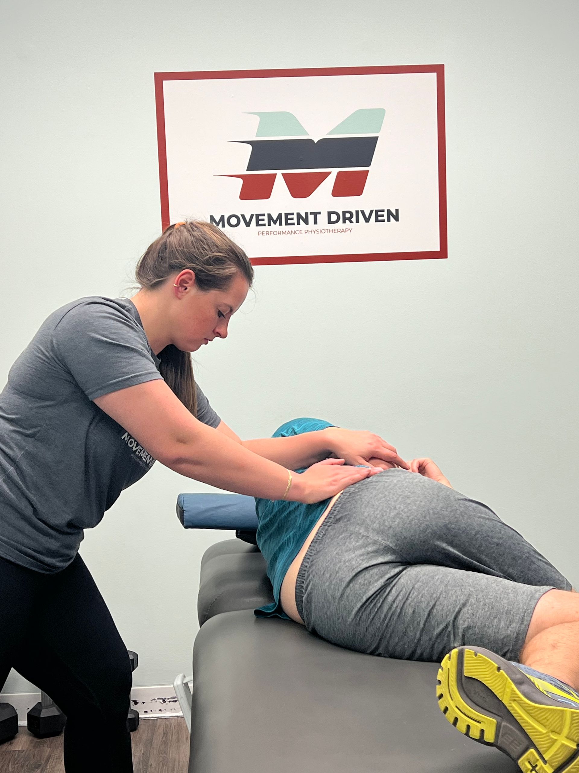 Therapist massaging a person's lower back while they lie on a treatment table. Movement Driven sign on wall.