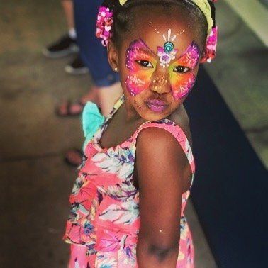 Child with colorful butterfly face paint and floral dress, looking over shoulder indoors.