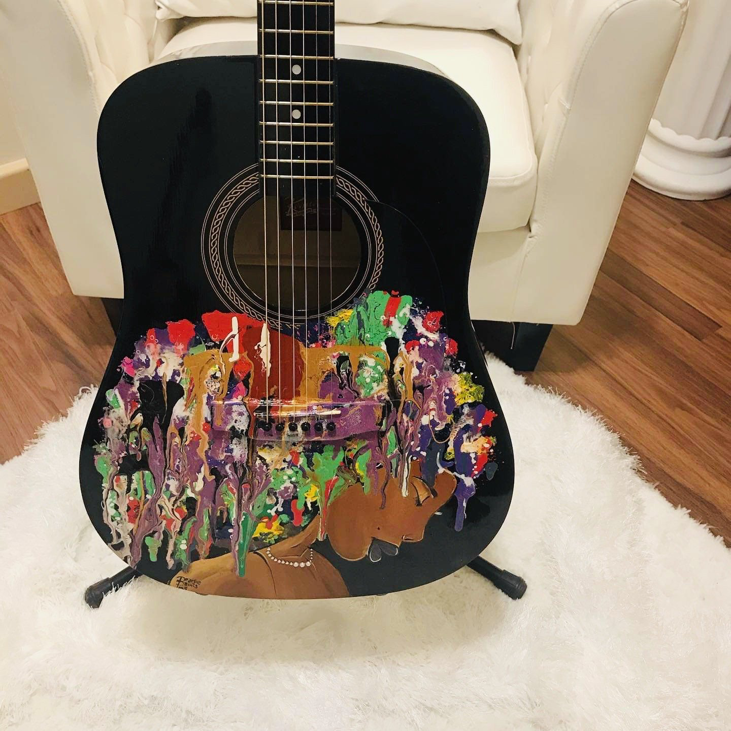 Black acoustic guitar with a colorful painted lower body, leaning on a white chair in a living room.