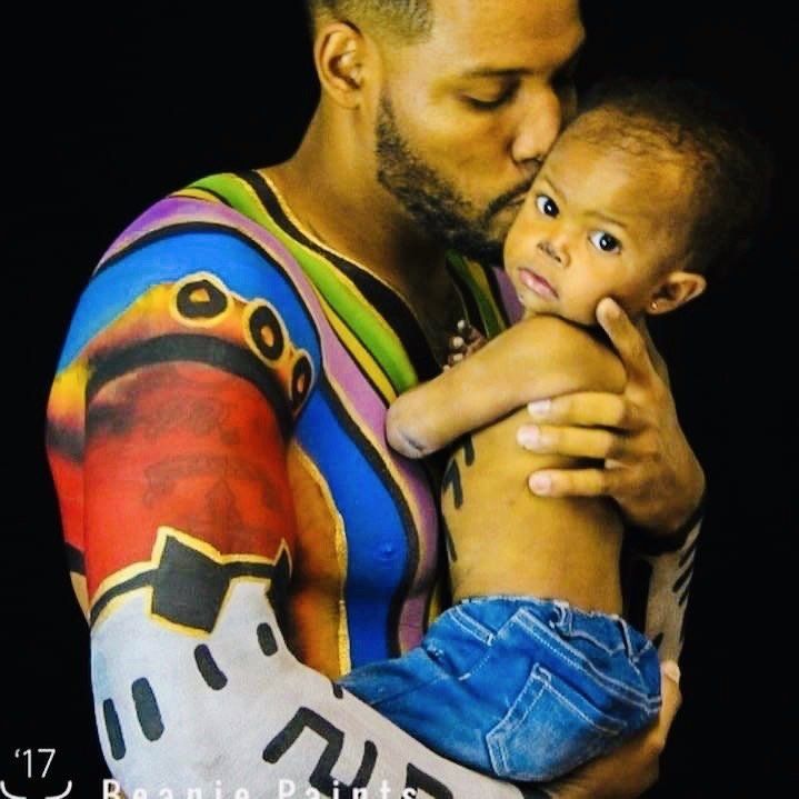 Adult holding and kissing a child with a colorful tattoo sleeve, both wearing blue jeans.