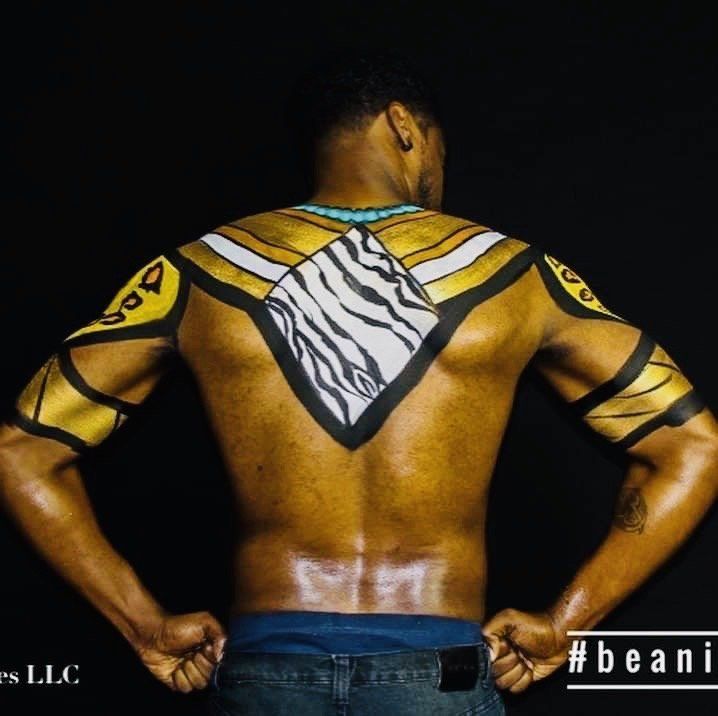 Back view of a person with yellow and black body paint, hands on hips, against a dark background