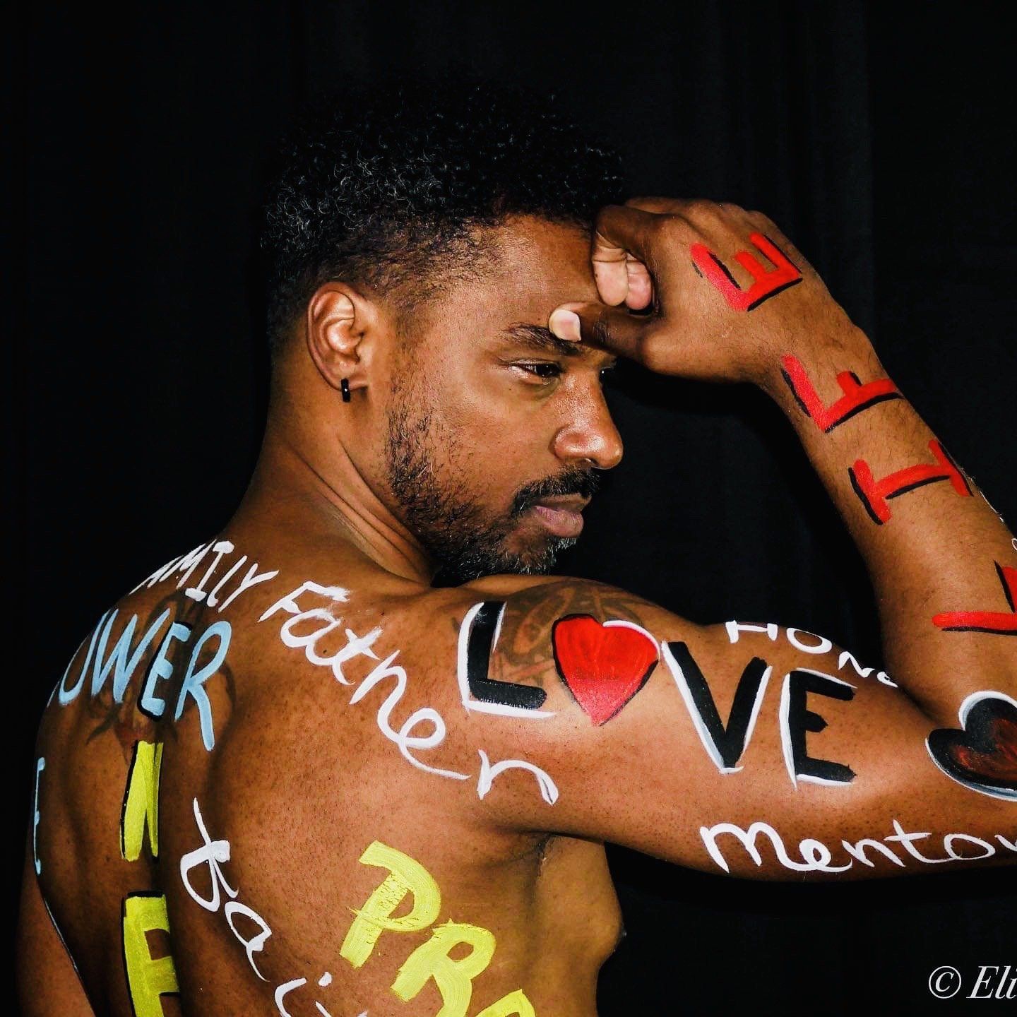 Man posing shirtless against a black background with colorful body paint text on arm and back.