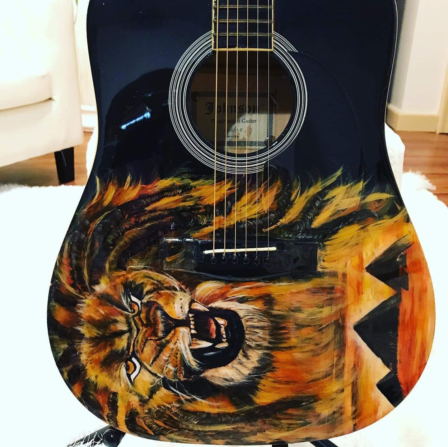 Acoustic guitar with a painted lion face and sunset scene on the body, standing upright indoors