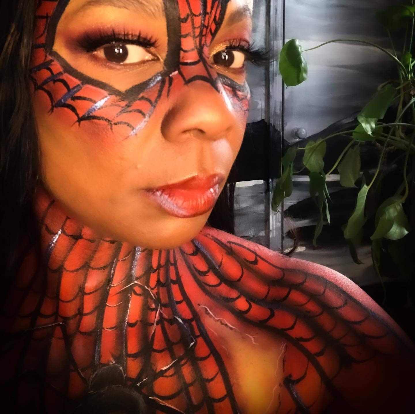 Close-up portrait with red-and-black spiderweb face and shoulder paint, leafy indoor background