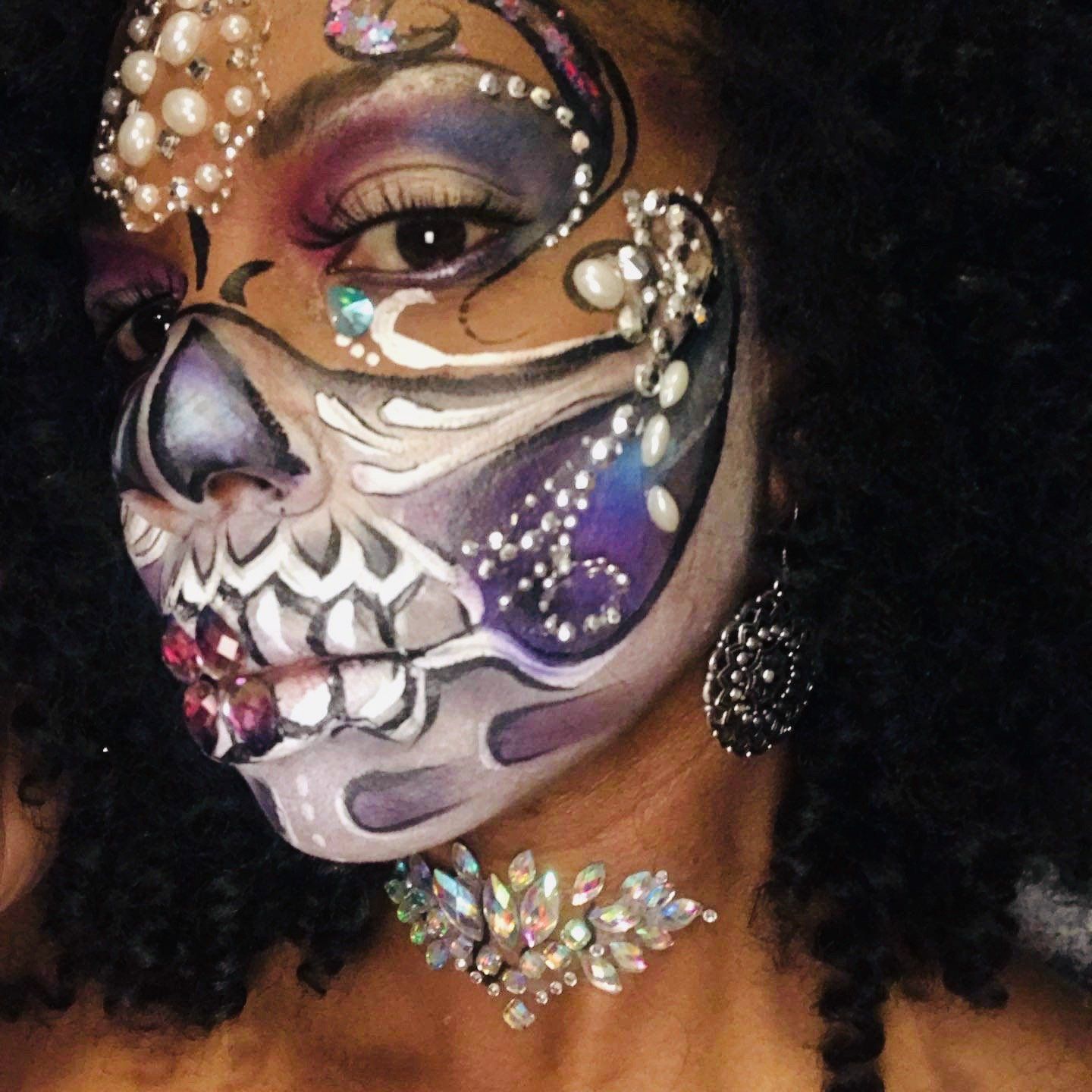 Face painted as a jeweled purple-and-white sugar skull with glitter and pearls on a black background