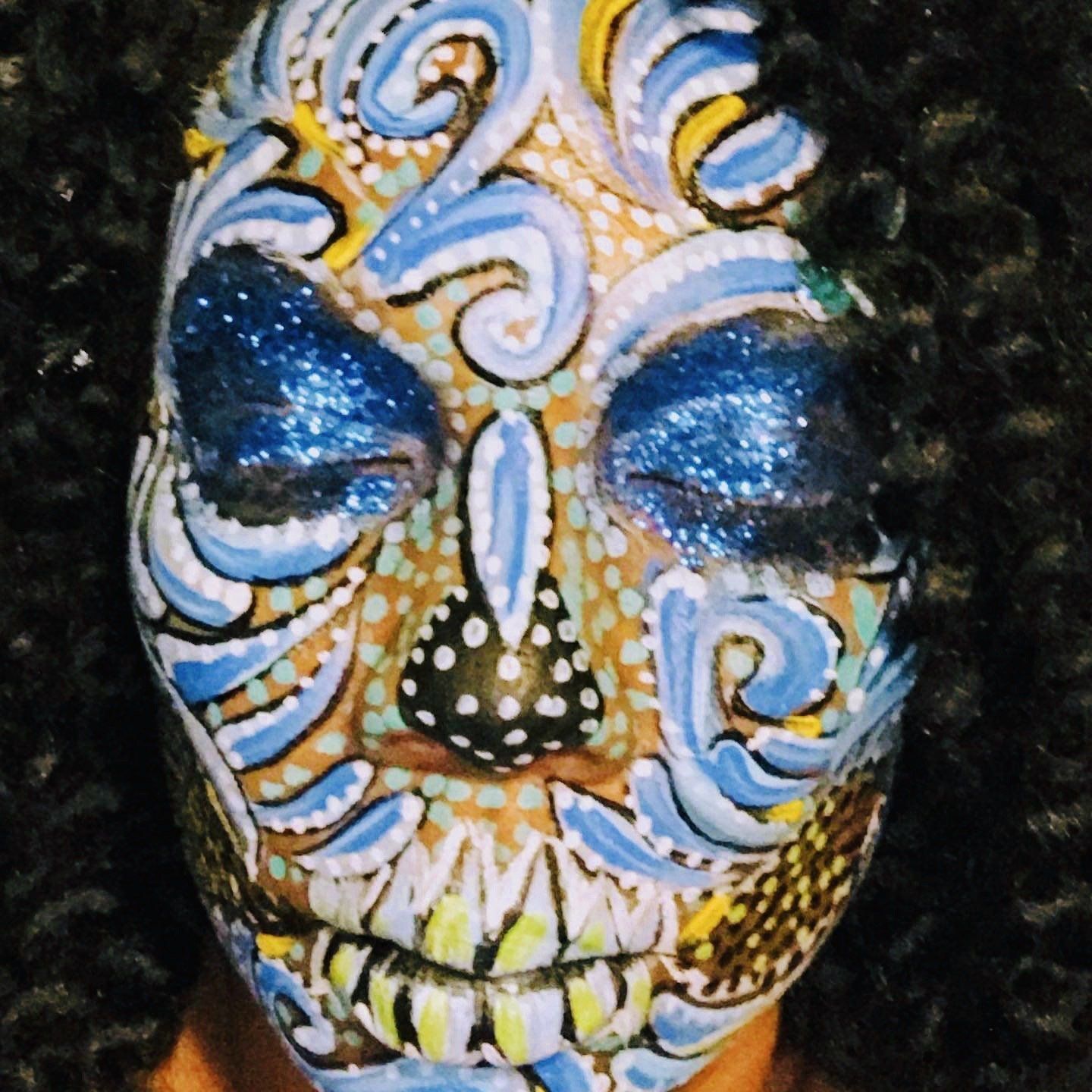 Face painted with intricate blue, white, and gold skull-like designs and glittery blue eye makeup