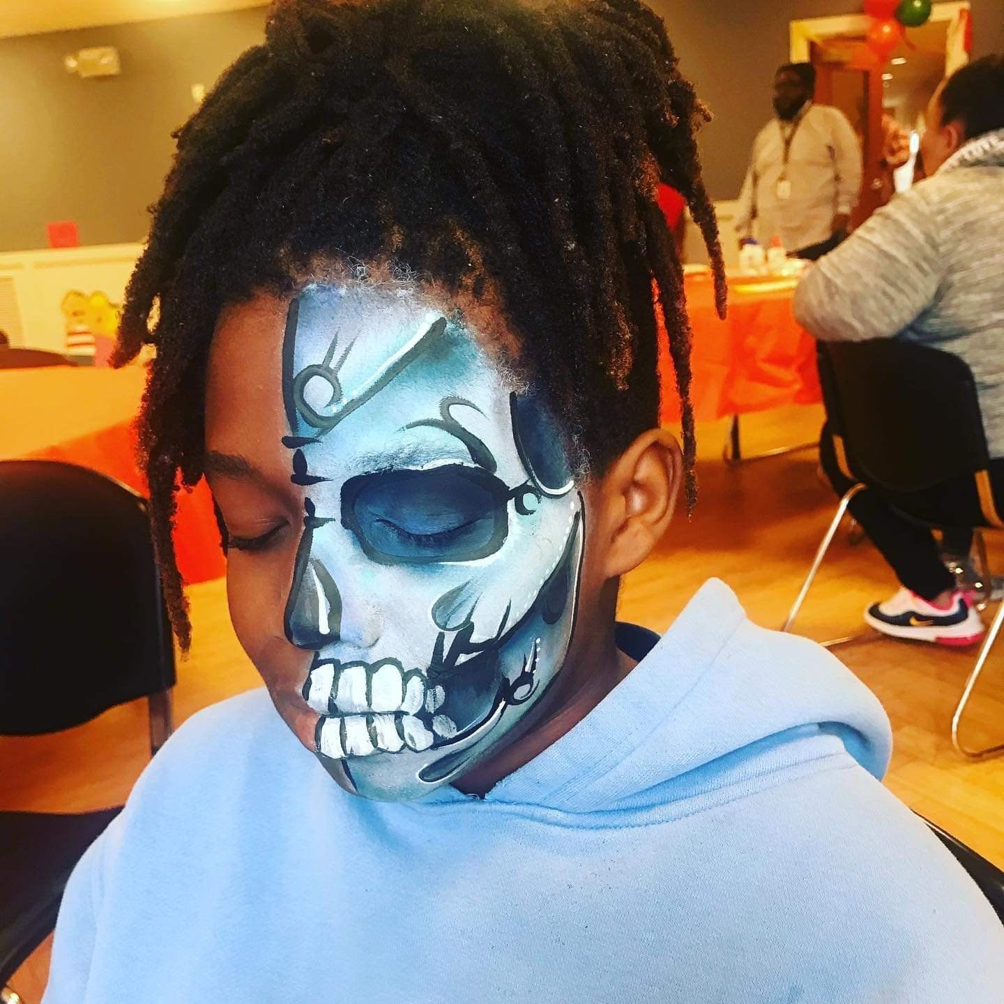 Child with skull face paint in a light blue hoodie, sitting indoors at a party.