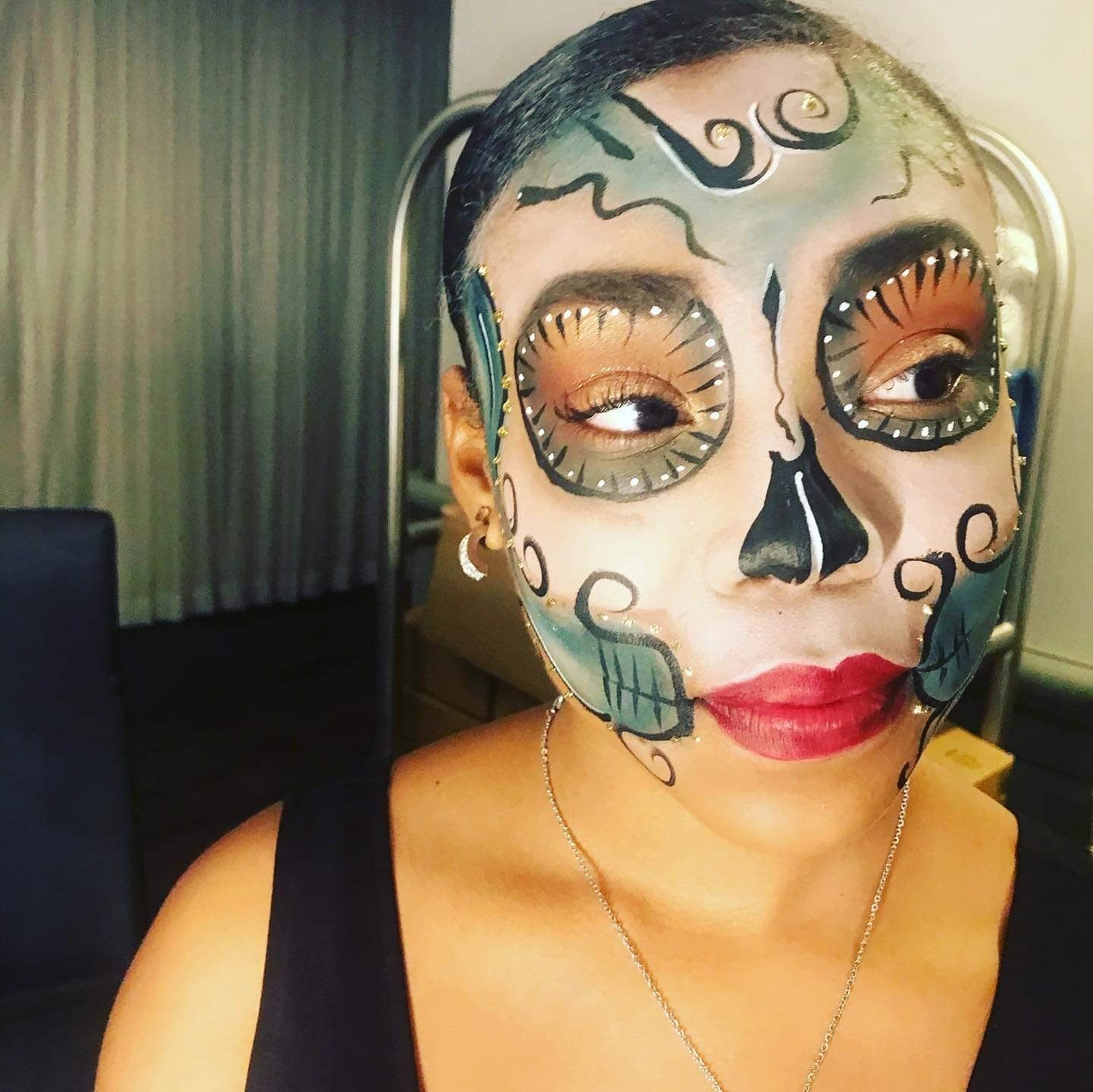 Person in a black top with colorful skull face paint and curled designs, looking sideways indoors