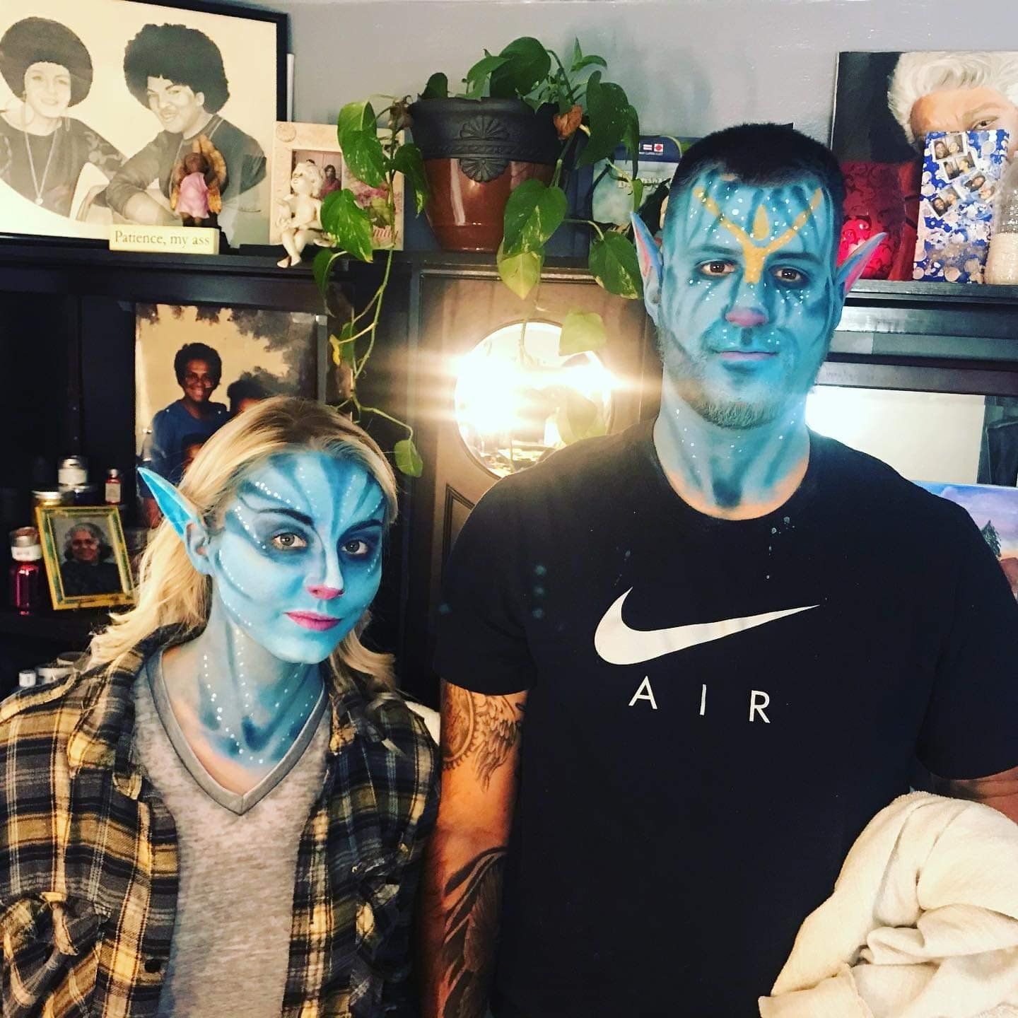 Two people in blue face paint posing indoors with shelves, plants, and framed photos behind them