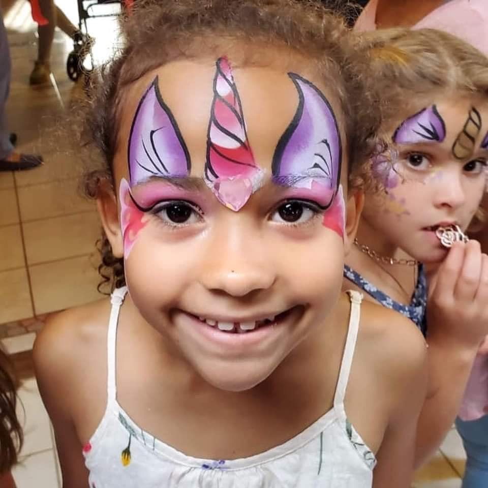 Smiling child with purple and pink face paint, with another face-painted child in the background