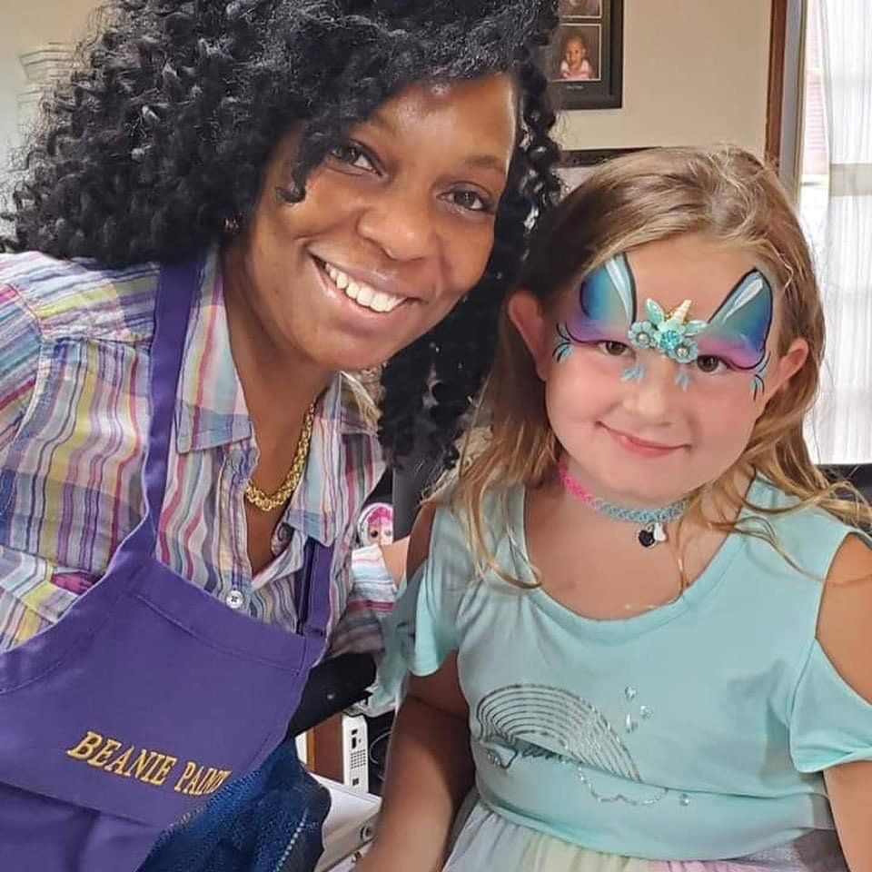 Two smiling people pose together, one wearing a purple apron and the other butterfly face paint.