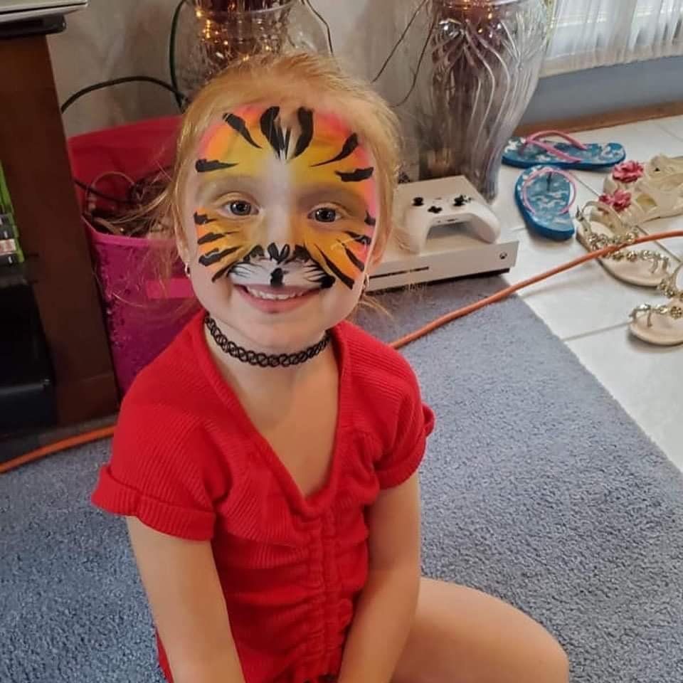 Child in a red outfit with tiger face paint, smiling indoors on a carpet.