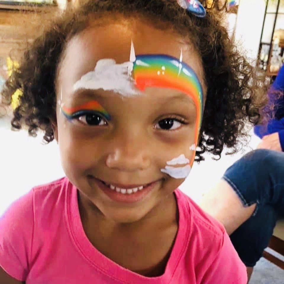 Smiling child with rainbow face paint and white cloud accents, wearing a pink shirt.