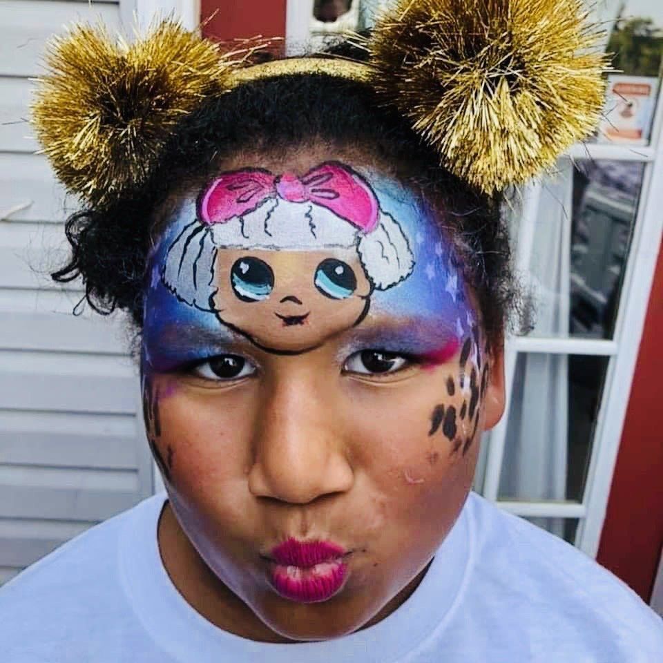 Child in festive face paint with a cartoon character on forehead and gold pom-pom headband, making a kissy face