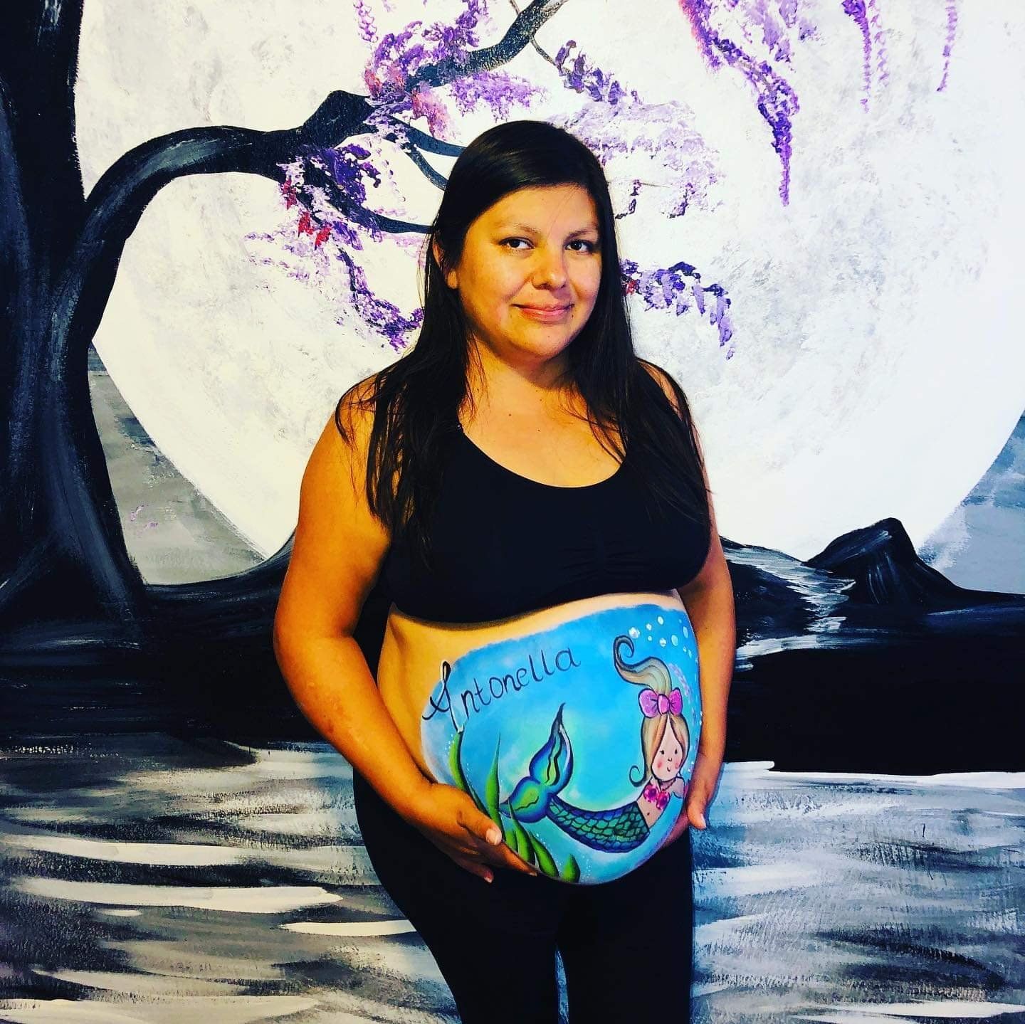 Pregnant woman in black top and jeans posing before a painted wall with a mermaid belly design