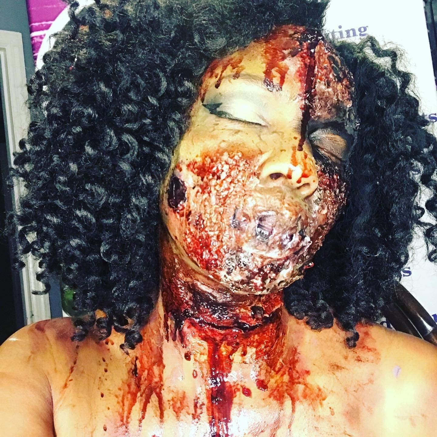 Person with dramatic blood-smeared makeup, eyes closed, wearing a curly black wig against a plain background