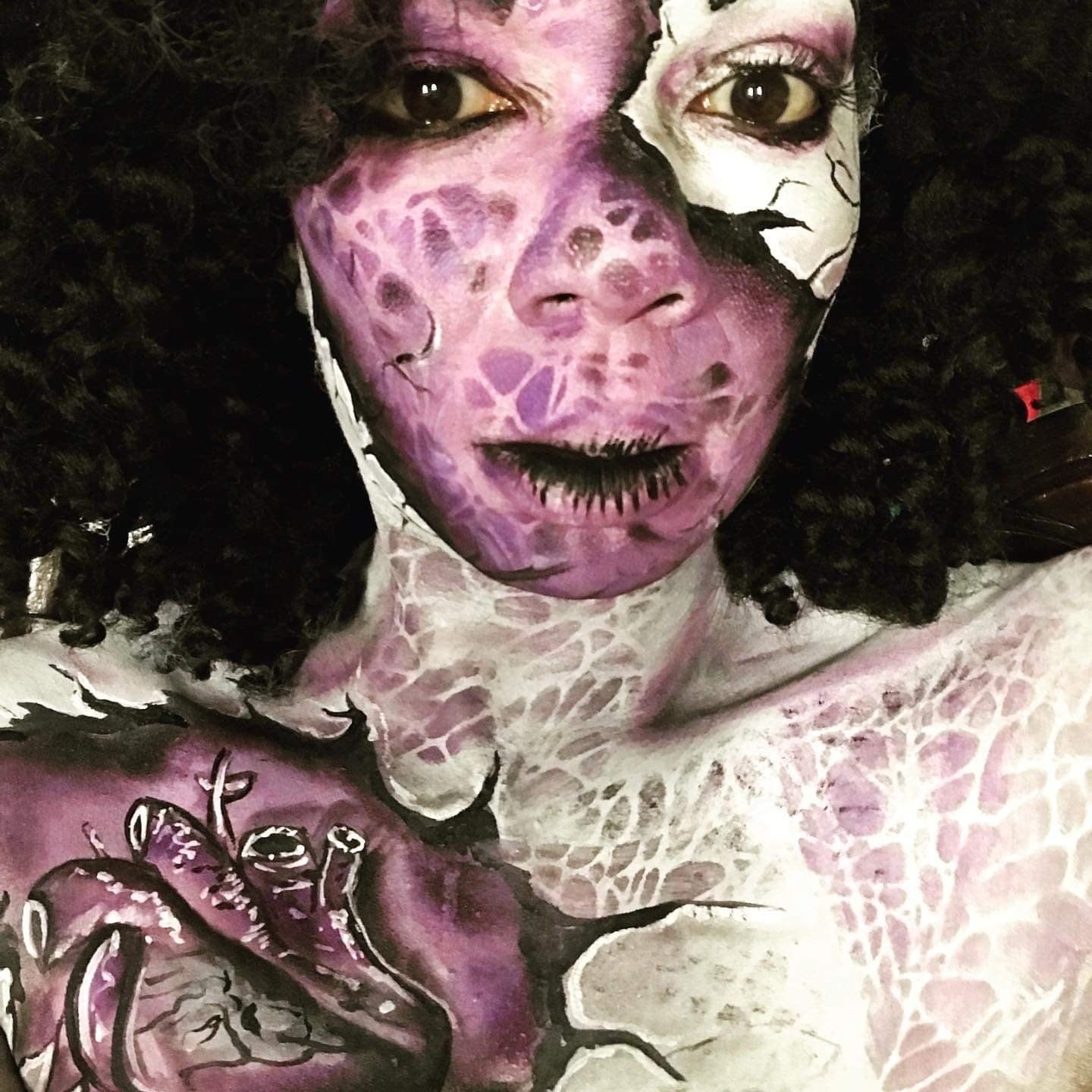 Close-up portrait of a person with purple and white reptile-like face and body paint, curly dark hair, and a raised painted hand