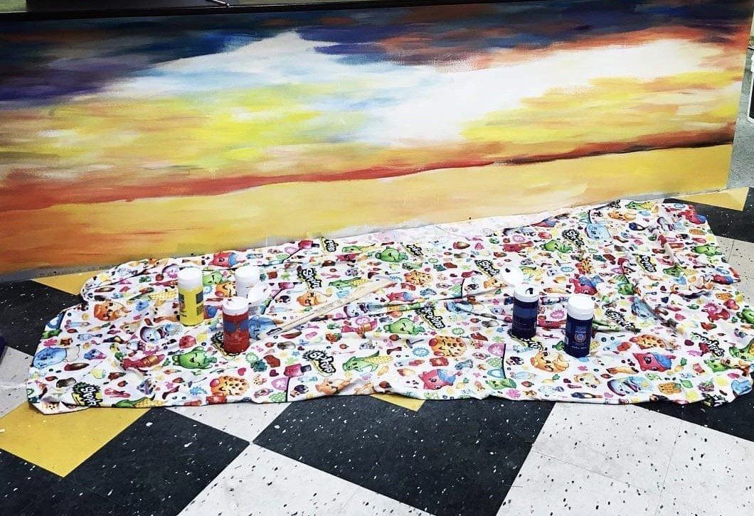 Colorful picnic blankets on a black-and-white checkered floor before a sunset mural, with cups and drinks on top