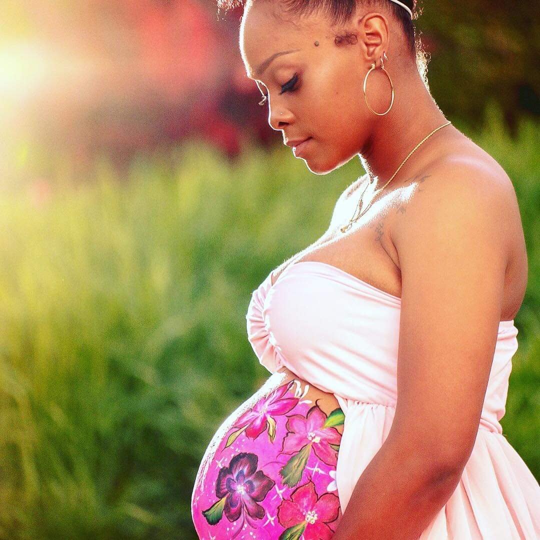 Pregnant woman in a white dress standing outdoors in a green garden, gently looking down at her belly