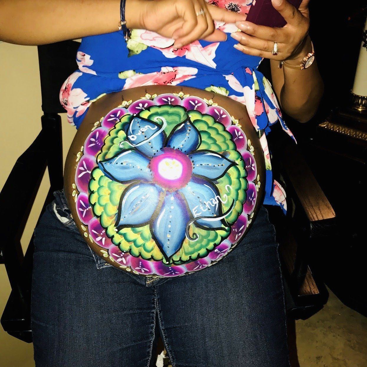 Person holding a colorful mandala-painted drum on their lap, wearing a blue floral top and jeans.