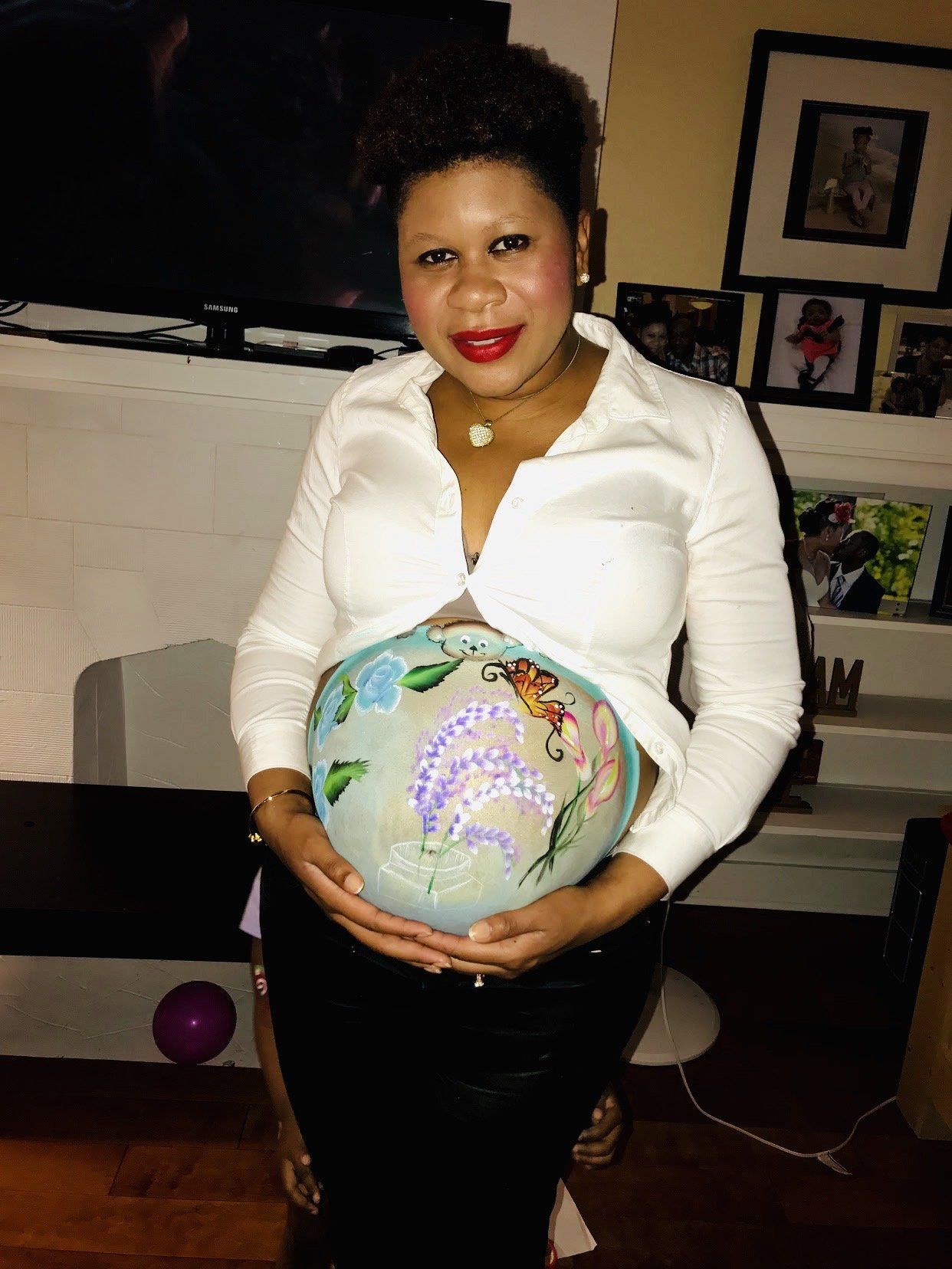 Person standing indoors, holding a decorated globe over their pregnant belly, wearing a white blouse and black skirt.