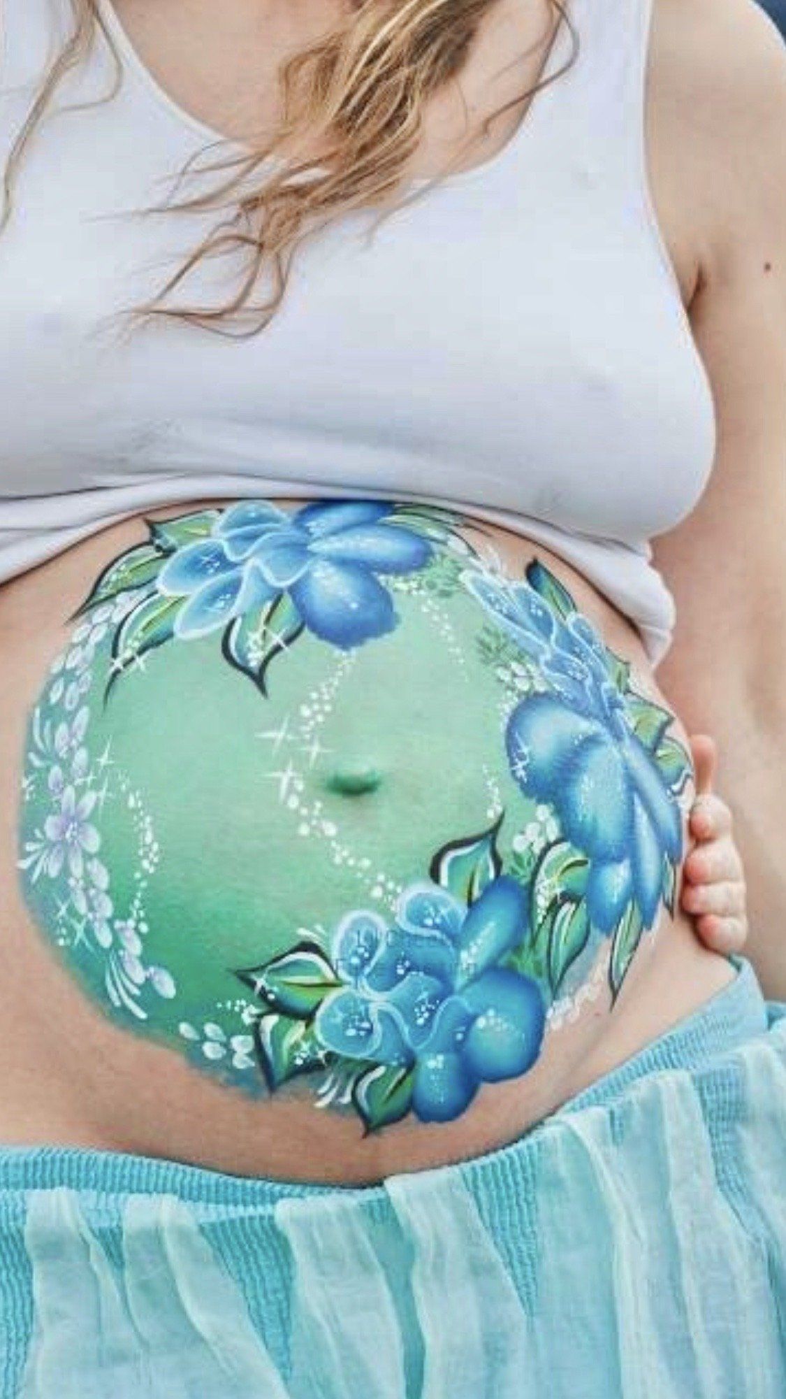 Painted belly with blue flowers on a pregnant abdomen, framed by a white top and blue skirt