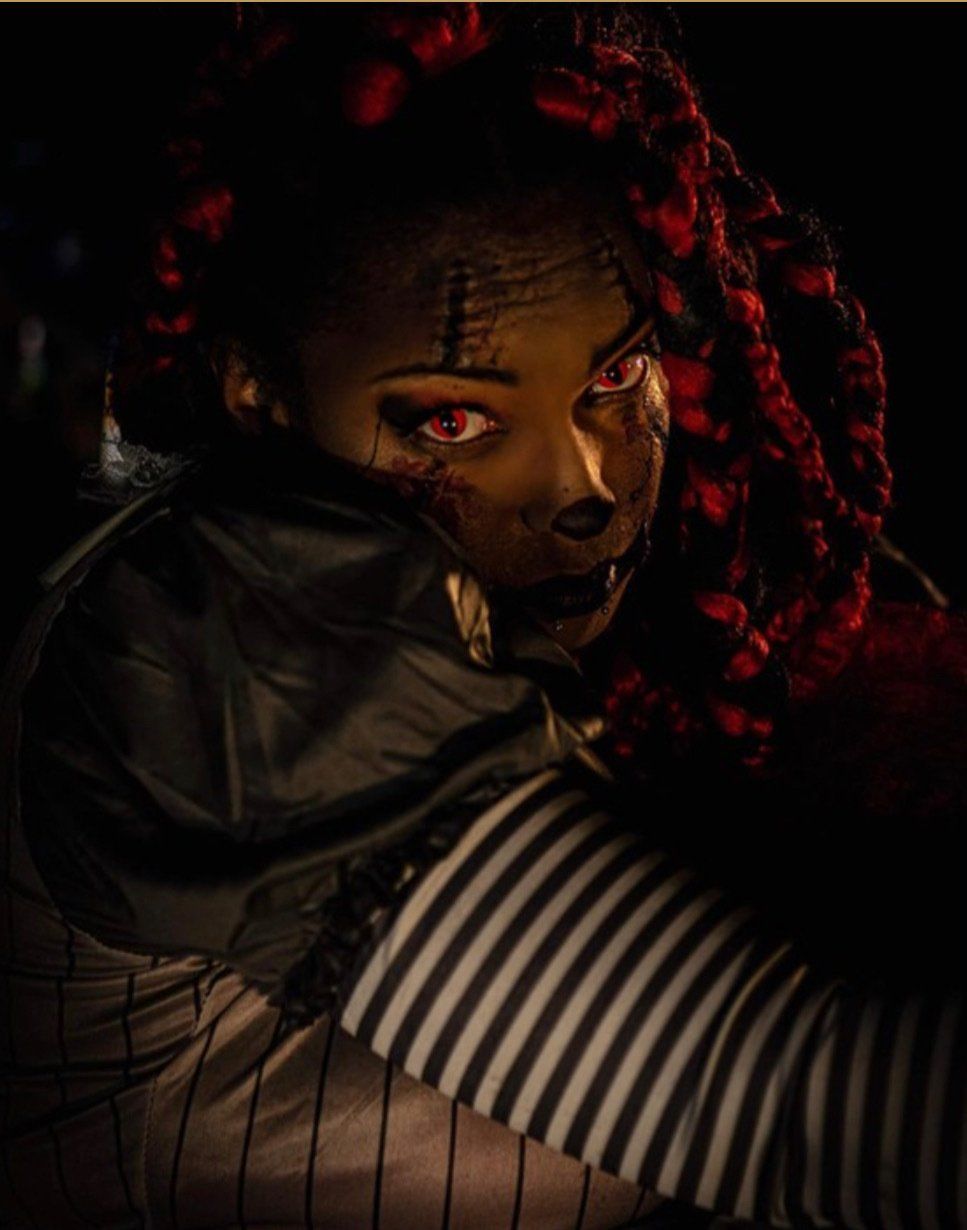 Close-up of a person with red braided hair in dim light, wearing striped sleeves and looking at the camera.